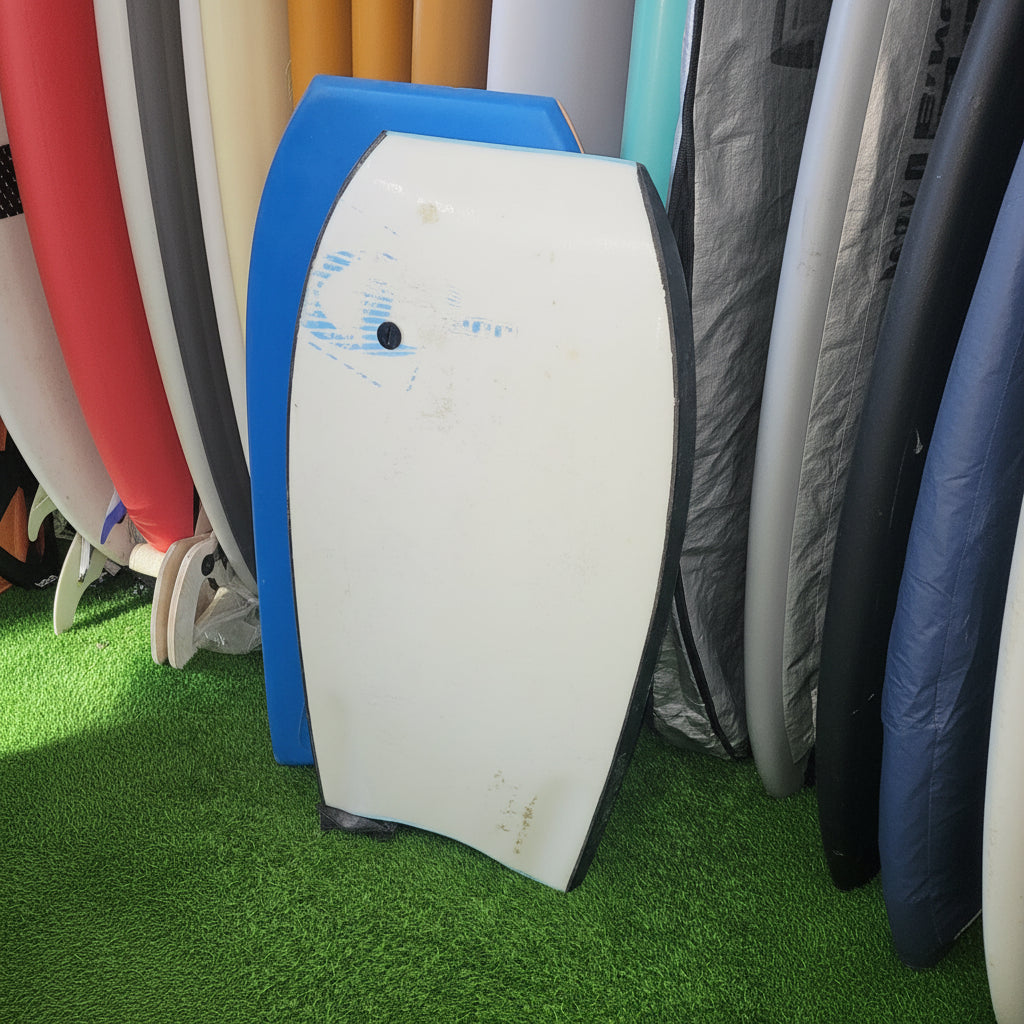 A white surfboard with a black nose and blue base, shown on artificial green turf next to other surfboards or boards.