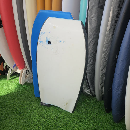 A white surfboard with a black nose and blue base, shown on artificial green turf next to other surfboards or boards.