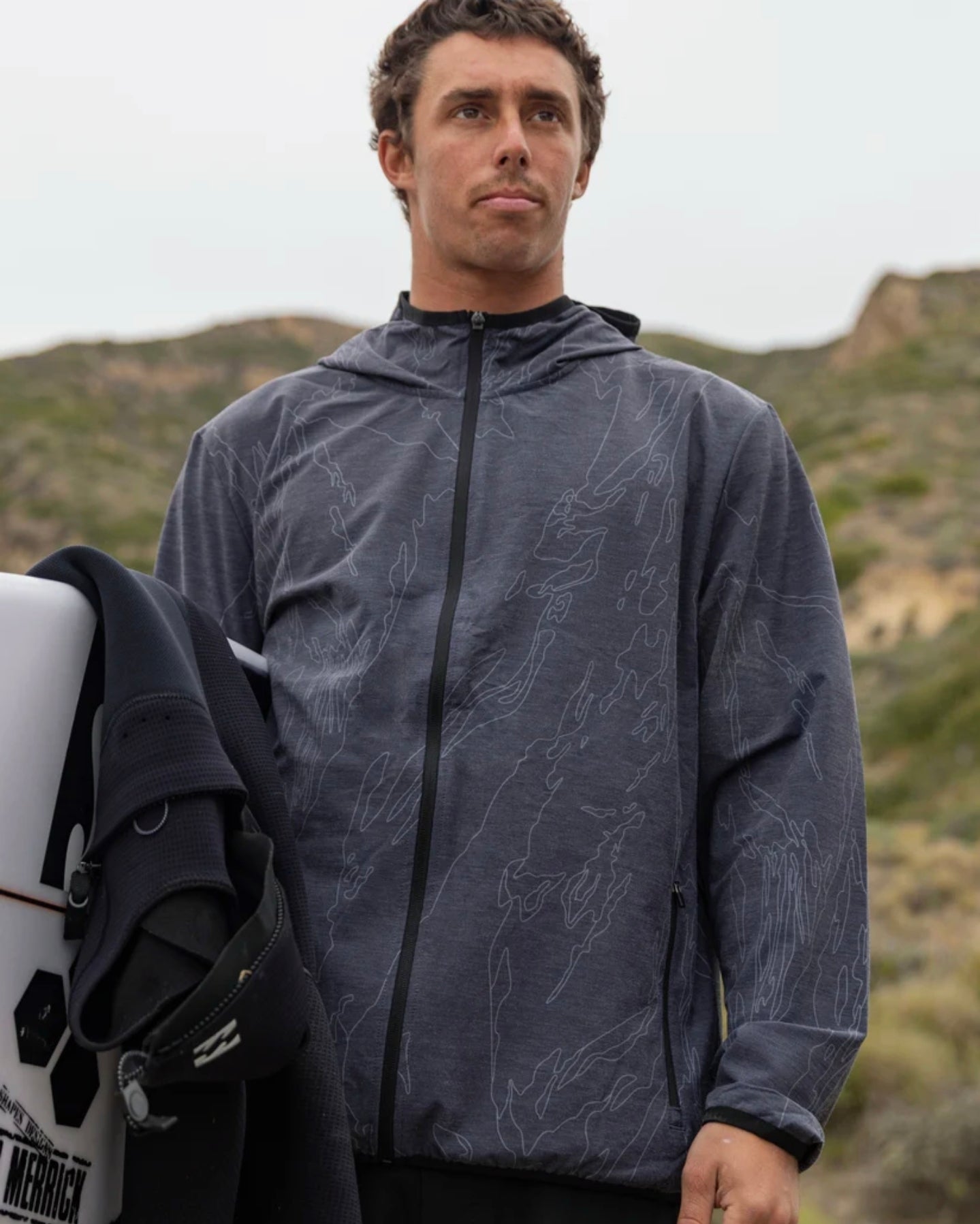 Man wearing a patterned jacket holding a surfboard with mountains in the background