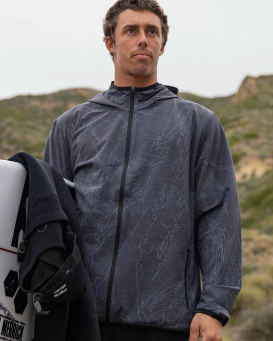 Man wearing a patterned jacket holding a surfboard with mountains in the background