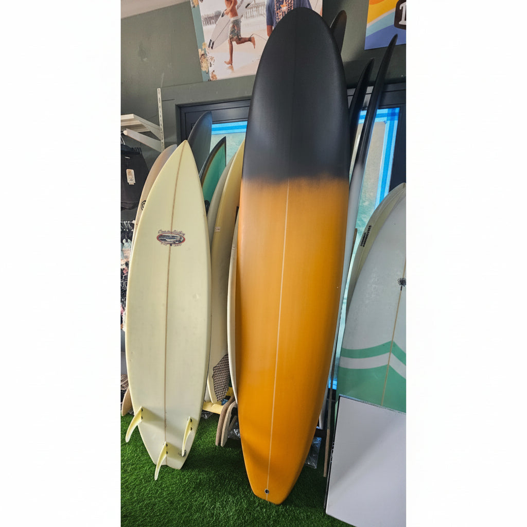 A long surfboard with a black-to-orange gradient finish stands upright in a shop, surrounded by other surfboards on display stands.