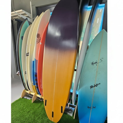 A stack of colorful surfboards displayed vertically on a stand, with a blue surfboard on top and a natural wood-grain board in the center, set against a background of green artificial turf.