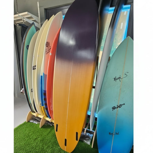 A stack of colorful surfboards displayed vertically on a stand, with a blue surfboard on top and a natural wood-grain board in the center, set against a background of green artificial turf.