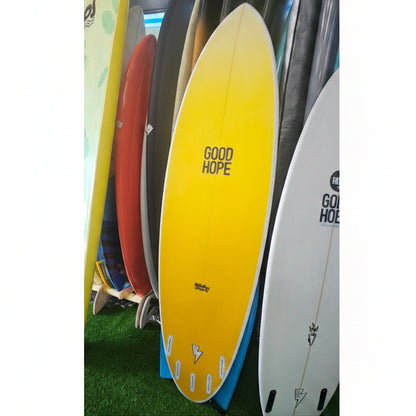 A yellow surfboard with 'GOOD HOPE' printed on the bottom, displayed vertically on a green artificial turf surface alongside other surfboards.