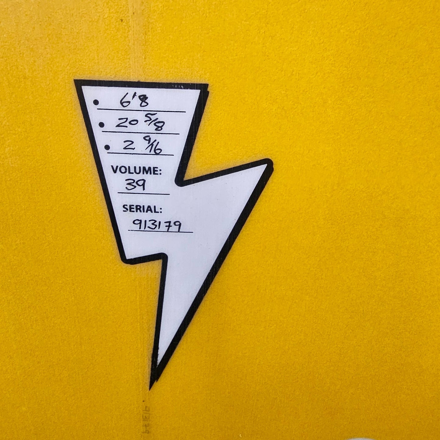 A close-up of a yellow surfboard or board surface with a white and black arrow-shaped sticker or label attached, plus two white plastic hardware pieces visible on the right side.
