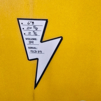 A close-up of a yellow surfboard or board surface with a white and black arrow-shaped sticker or label attached, plus two white plastic hardware pieces visible on the right side.