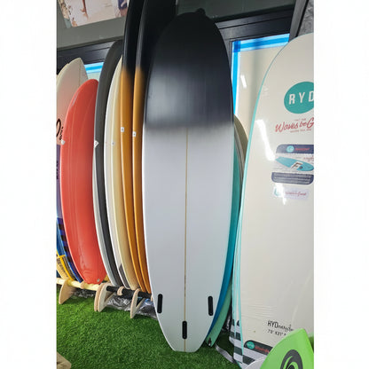 A stack of surfboards in various colors, including black, white, orange, and red, displayed on a stand in a store setting with artificial green turf visible in the background.