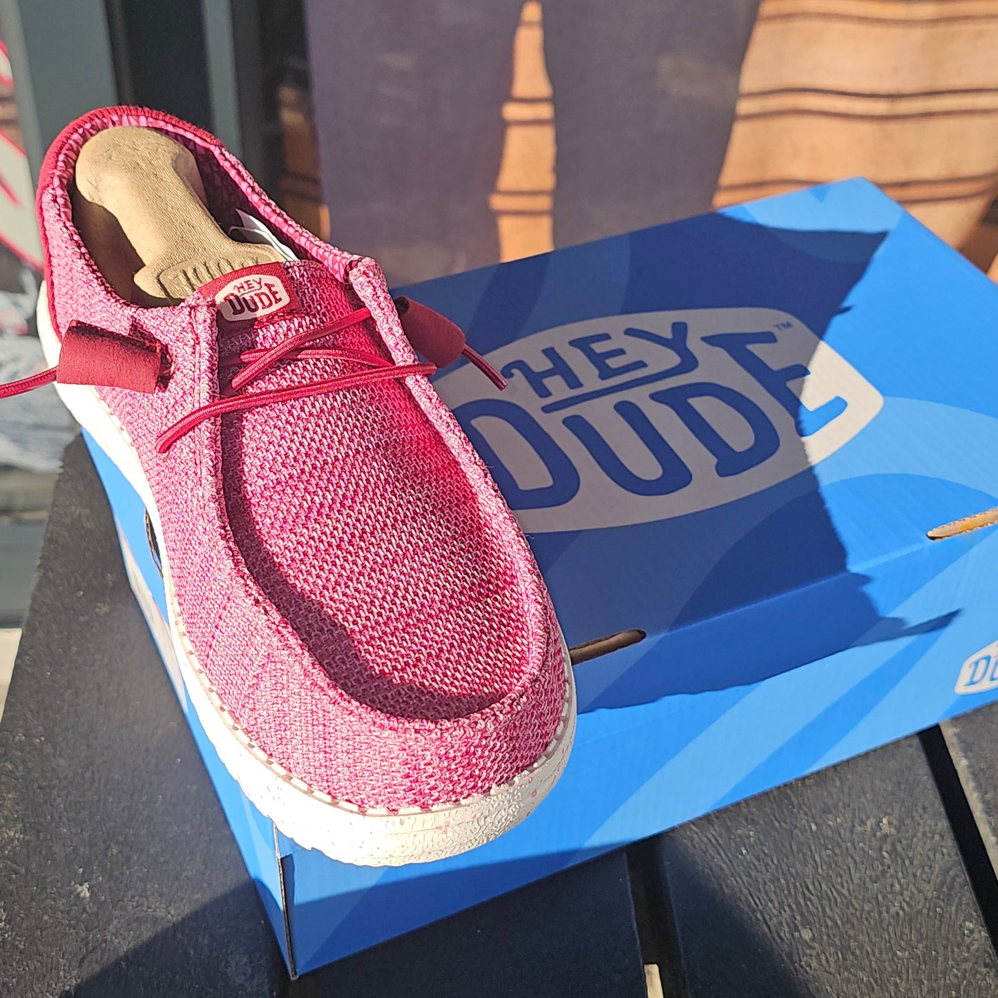 A pink knit slip-on shoe with a white sole, shown next to its blue box on a dark wooden surface.