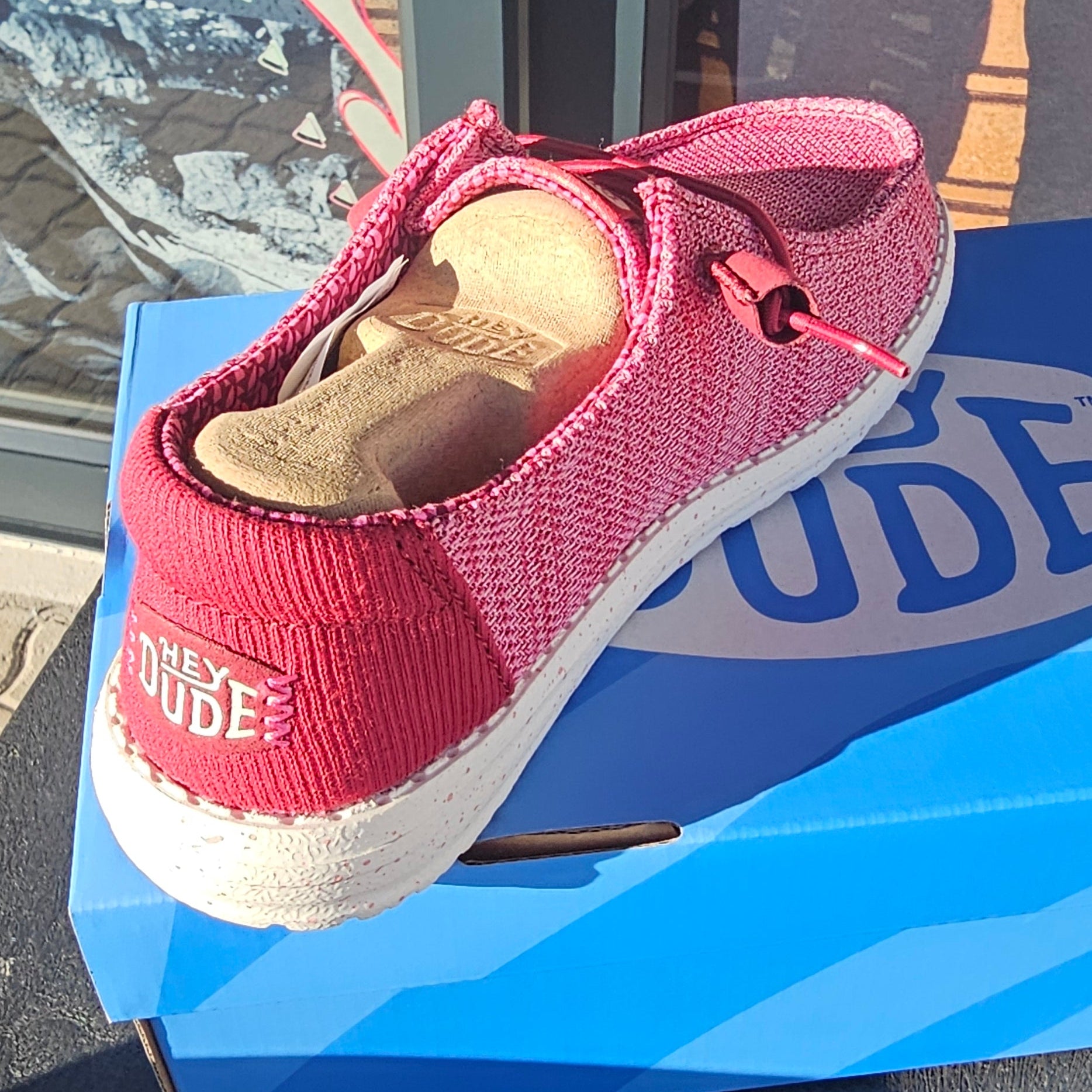 A pink mesh slip-on shoe with a white sole and a blue box underneath, photographed on a dark surface near a window.