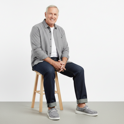 Man sitting on a stool wearing a gray shirt, navy pants, and gray sneakers against a white background
