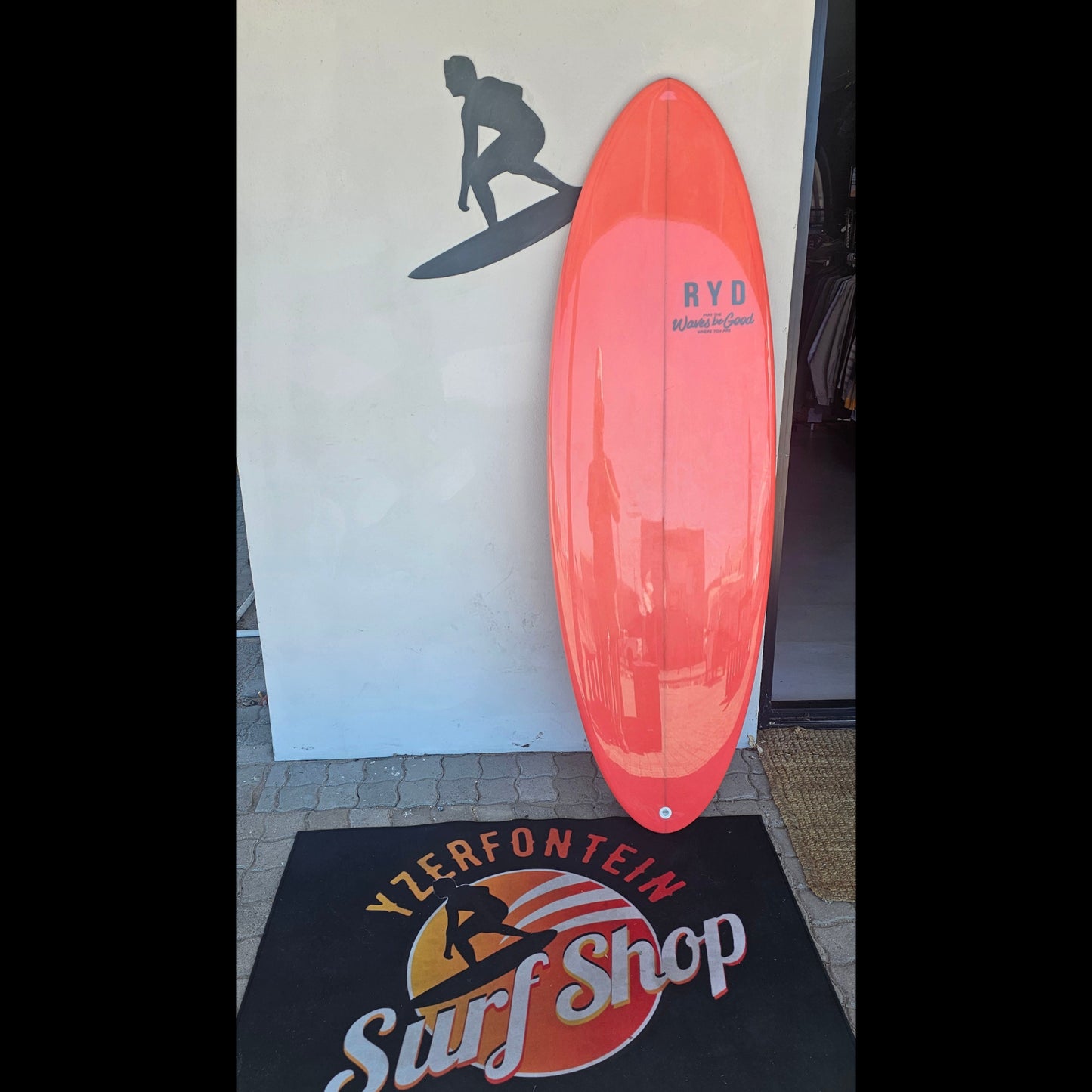 ligt red surfboard with 'RYD' branding on a mat in front of a wall with a silhouette of a surfer.