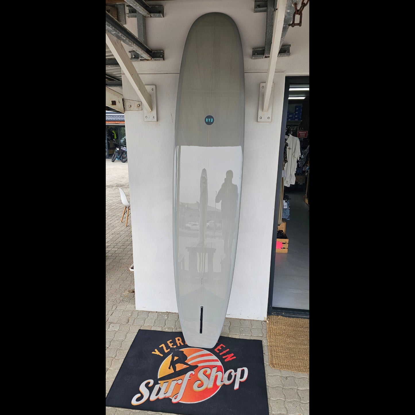 Longboard-style surfboard on a white stand with a surf shop sign in the background.