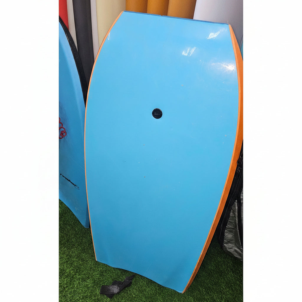 A light blue bodyboard with an orange edge, shown on a green artificial turf surface.
