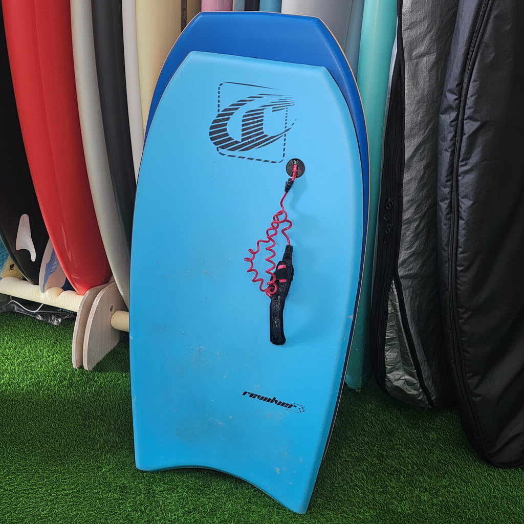 A light blue bodyboard with a black handle and red coiled leash, photographed on artificial green turf with other boards partially visible in the background.