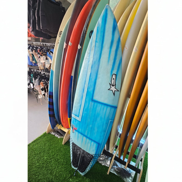 A blue surfboard with a white star logo stands upright on a display rack alongside other colorful surfboards, set against a store background with clothing racks and a green artificial turf floor.