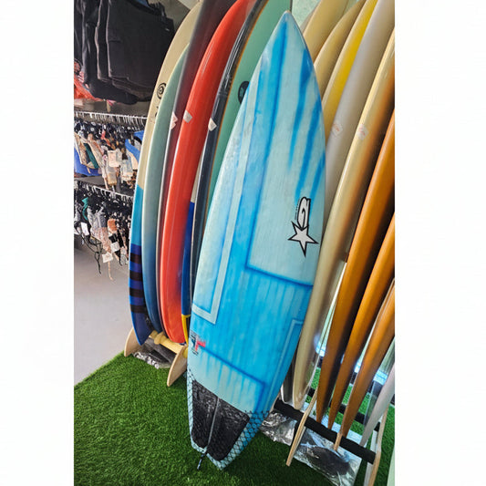 A blue surfboard with a white star logo stands upright on a display rack alongside other colorful surfboards, set against a store background with clothing racks and a green artificial turf floor.